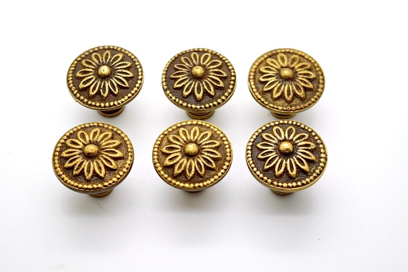 May include: Six gold-toned cabinet knobs with a floral design. Each knob features a raised, circular flower with a central dot.