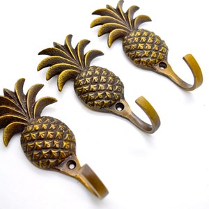 May include: Three bronze-colored pineapple-shaped wall hooks. Each hook features a detailed pineapple design with a curved hook extending from the base. The hooks are set against a white background, highlighting their decorative and functional nature.