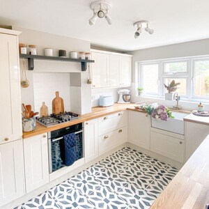 May include: A white kitchen with white cabinets, wood countertops, and a black and white patterned tile floor. The kitchen features a white farmhouse sink, a gas stove, and a black oven.