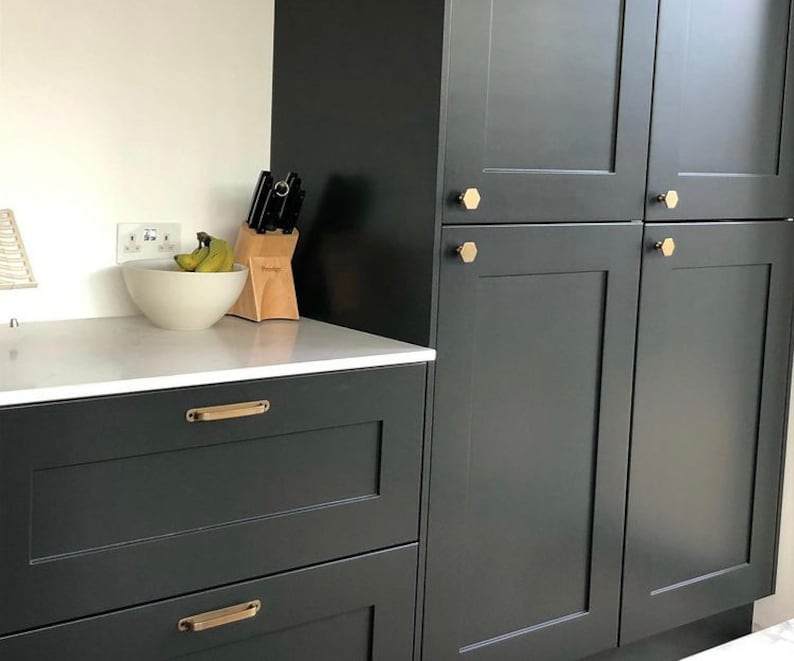 May include: A dark green kitchen cabinet with two drawers and a set of upper cabinets. The drawers have brushed gold handles. The upper cabinets have hexagonal brushed gold knobs. The cabinet is in a modern kitchen.