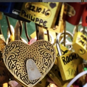 May include: A close-up of a heart-shaped, gold-colored padlock with an ornate design. The padlock is in focus, surrounded by other padlocks of various colors and shapes, some with handwritten names.