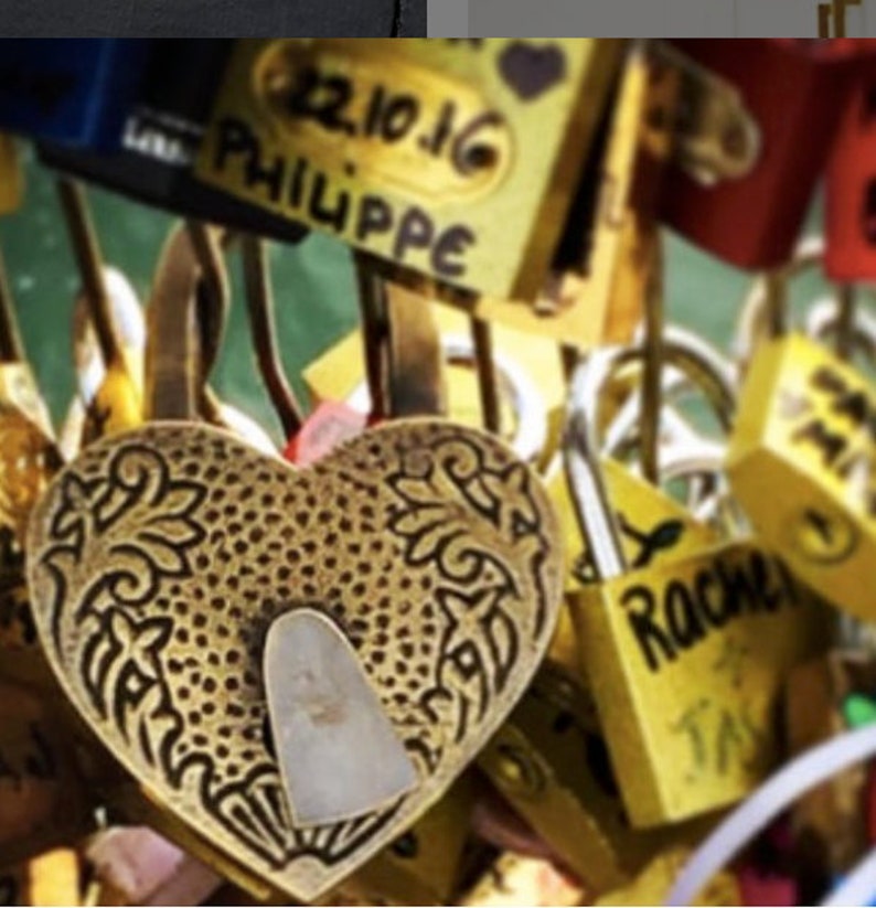 May include: A close-up of a heart-shaped, gold-colored padlock with an ornate design. The padlock is in focus, surrounded by other padlocks of various colors and shapes, some with handwritten names.