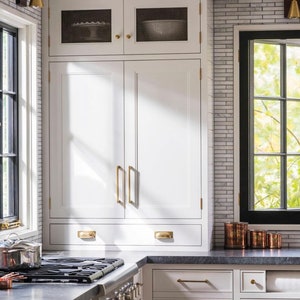 May include: White kitchen cabinets with brass hardware. The cabinets have a built-in design with a drawer and upper cabinets. A stainless steel stove and a window with a view of trees are also visible.