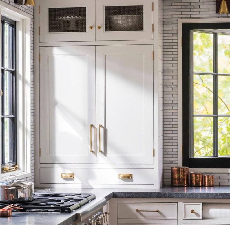 May include: White kitchen cabinets with brass hardware. The cabinets have a built-in design with a drawer and upper cabinets. A stainless steel stove and a window with a view of trees are also visible.