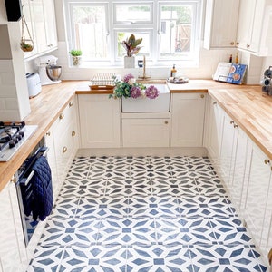May include: A white kitchen with wood countertops and white cabinets. The floor is tiled with a black and white geometric pattern. There is a window with a view of a garden.