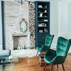 May include: A cozy living room scene featuring a fireplace with a gold-framed mirror, flanked by a stack of firewood and a dark blue bookshelf. Two emerald green velvet armchairs with wooden legs sit on a wooden floor, creating a stylish and inviting space.