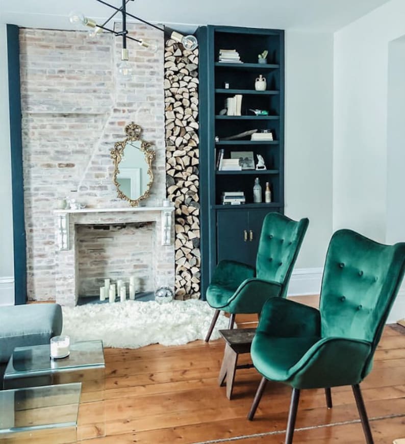 May include: A cozy living room scene featuring a fireplace with a gold-framed mirror, flanked by a stack of firewood and a dark blue bookshelf. Two emerald green velvet armchairs with wooden legs sit on a wooden floor, creating a stylish and inviting space.