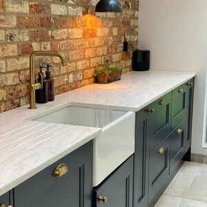 May include: A kitchen with a white countertop, a white farmhouse sink, and dark blue cabinets with brass hardware. The faucet is gold, and the backsplash is brick. There are brown soap dispensers and a small plant.