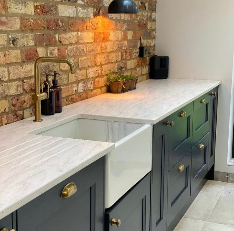 May include: A kitchen with a white countertop, a white farmhouse sink, and dark blue cabinets with brass hardware. The faucet is gold, and the backsplash is brick. There are brown soap dispensers and a small plant.