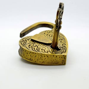 May include: A heart-shaped, antique brass padlock with an ornate floral design. The key is inserted in the lock. The lock is open. The padlock is on a white background.