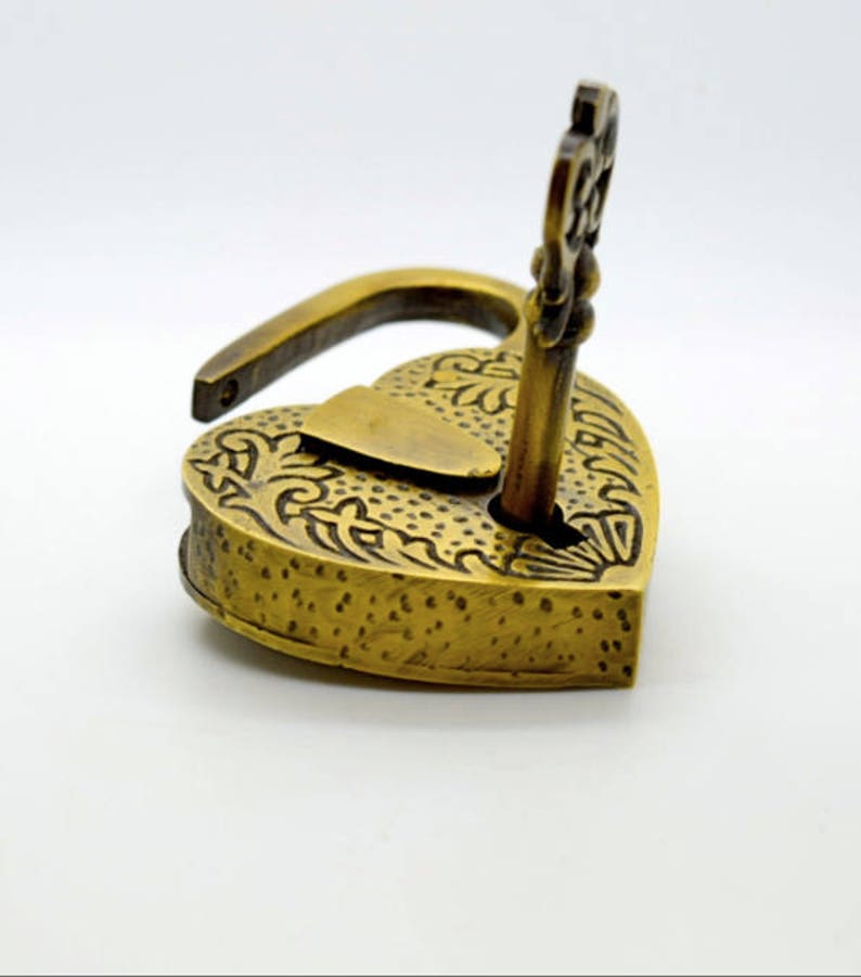 May include: A heart-shaped, antique brass padlock with an ornate floral design. The key is inserted in the lock. The lock is open. The padlock is on a white background.