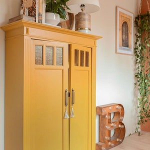 May include: A mustard yellow cabinet with glass panel doors and brass handles. The cabinet is topped with books, a lamp, and a potted plant. A framed picture and a decorative letter "B" are in the background.