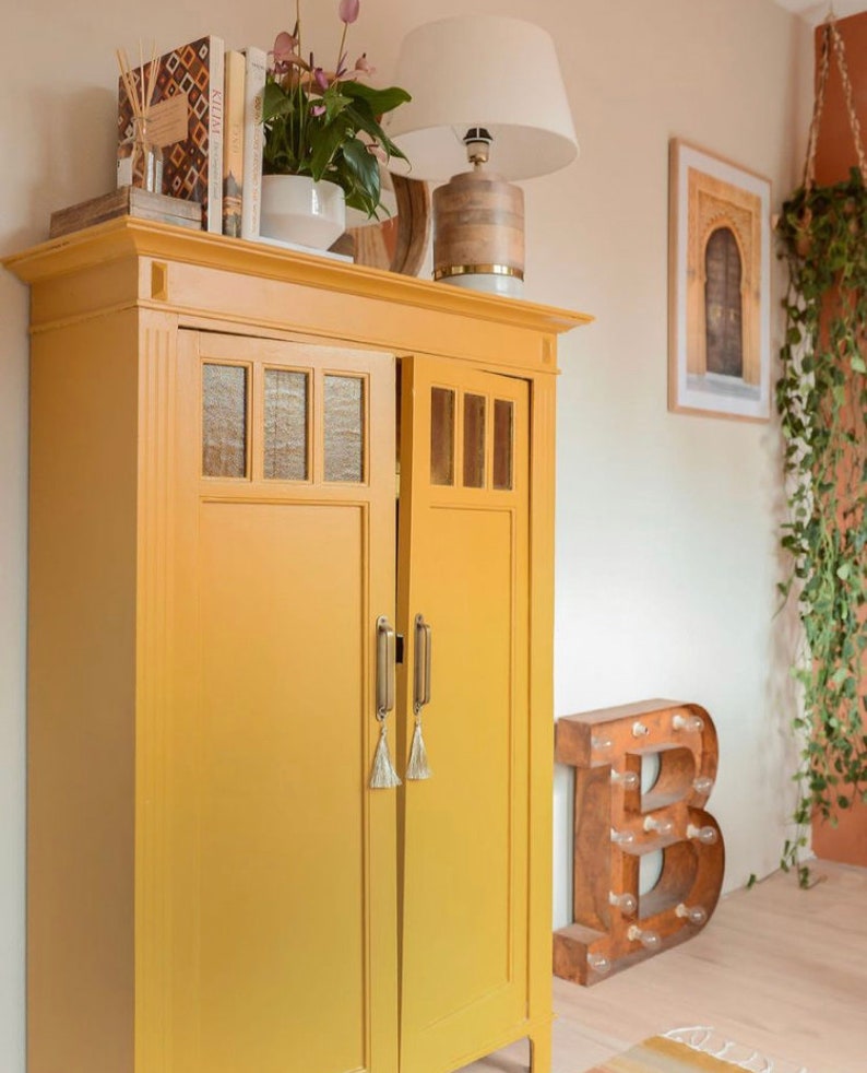 May include: A mustard yellow cabinet with glass panel doors and brass handles. The cabinet is topped with books, a lamp, and a potted plant. A framed picture and a decorative letter "B" are in the background.