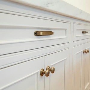 May include: White kitchen cabinets with brass hardware. The cabinets have a recessed panel design and are accented with rectangular and round brass pulls. A white countertop is visible above the cabinets.