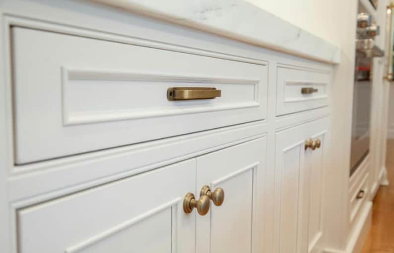 May include: White kitchen cabinets with brass hardware. The cabinets have a recessed panel design and are accented with rectangular and round brass pulls. A white countertop is visible above the cabinets.