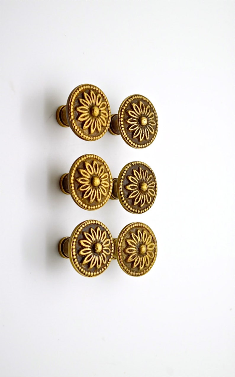 May include: Six gold-toned metal knobs with a floral design. Each knob has a round base and a raised floral pattern.