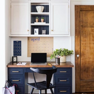 May include: A home office setup featuring a built-in desk with navy blue drawers and a wooden countertop. Above, white cabinets with open shelving display decor. A black chair sits in front of the desk, with a laptop and calendar visible.