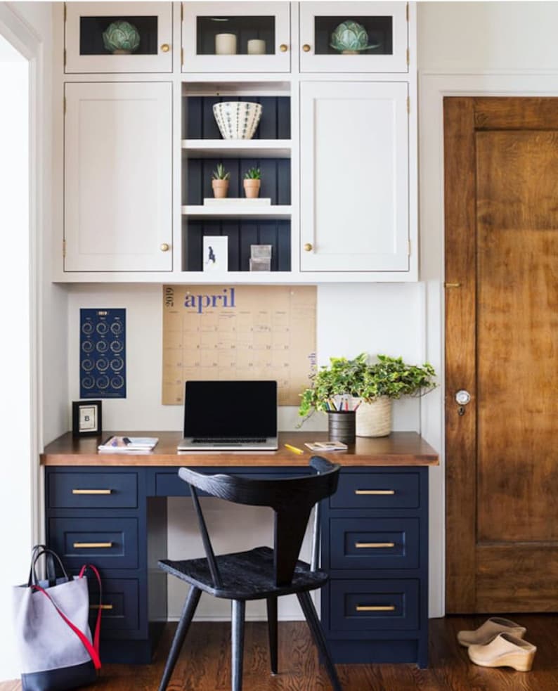 May include: A home office setup featuring a built-in desk with navy blue drawers and a wooden countertop. Above, white cabinets with open shelving display decor. A black chair sits in front of the desk, with a laptop and calendar visible.