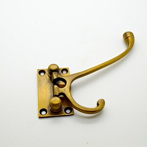 May include: A brass-colored wall hook with a rectangular base and a curved hook. The base has four screw holes and two decorative knobs. The hook extends outward and curves downward, suitable for hanging items. The overall design is vintage.