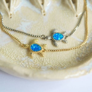 Puede incluir: Dos cadenas de oro con un dije de tortuga de ópalo azul en cada una. Las cadenas están sobre un plato de cerámica blanca.