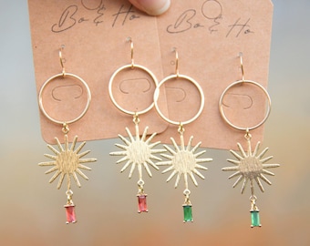 May include: Gold hoop earrings with a dangling sunburst charm and a small red or green gemstone. The earrings are displayed on a brown card with the text "Bo & Ho" written on it.