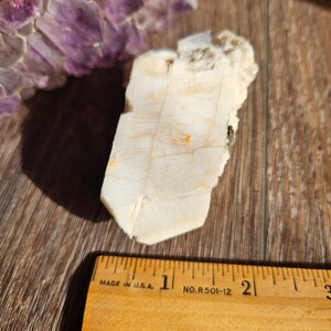 May include: A white, translucent crystal with a rough, textured surface. The crystal is about 2 inches long and is sitting on a wooden surface next to a ruler.