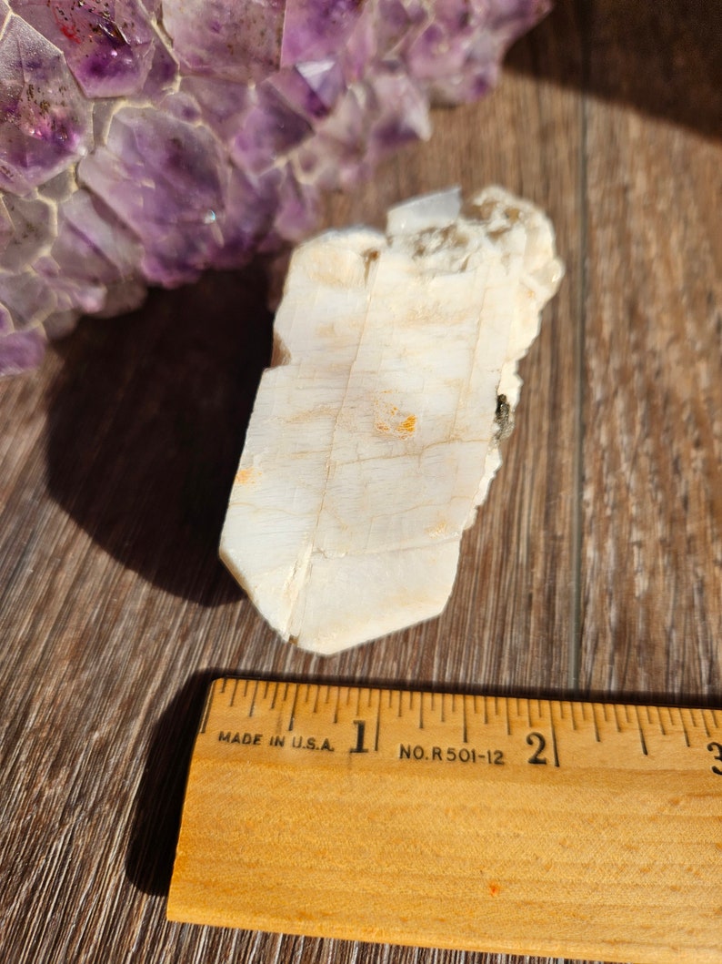 May include: A white, translucent crystal with a rough, textured surface. The crystal is about 2 inches long and is sitting on a wooden surface next to a ruler.