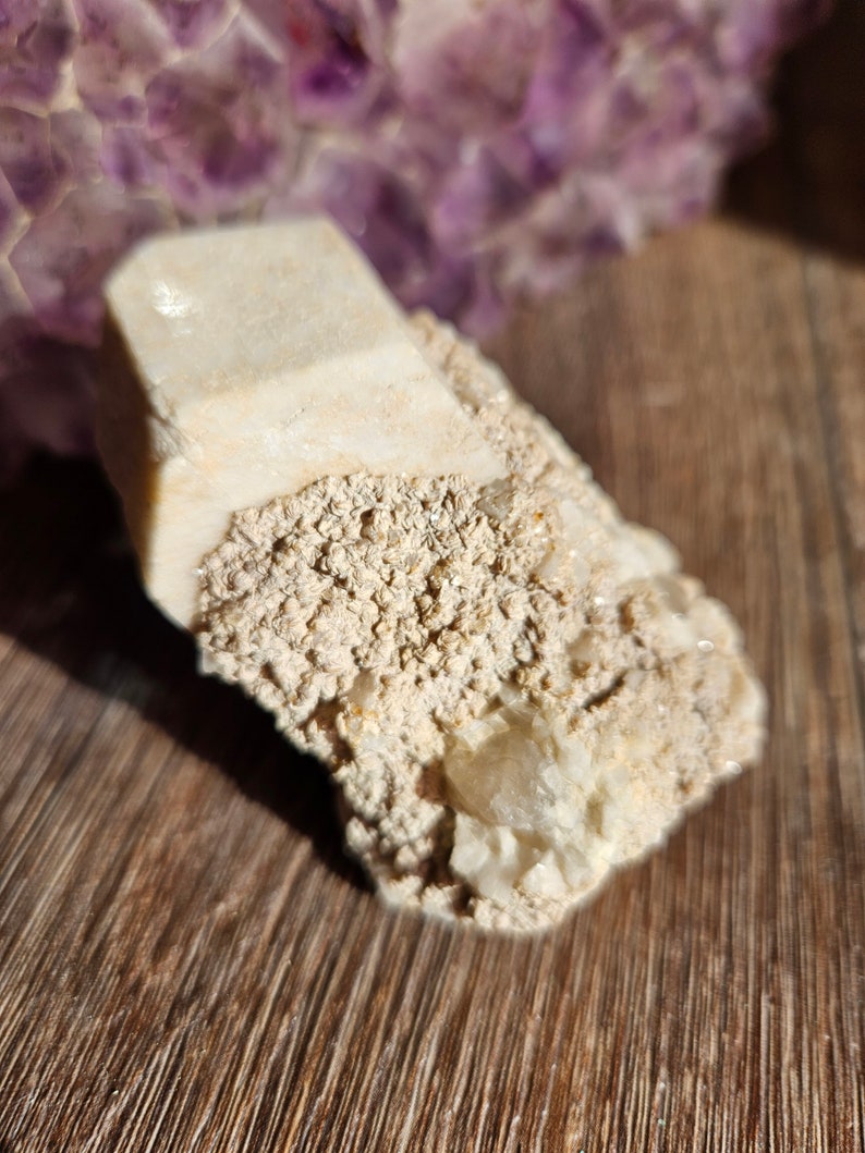 May include: A white crystal cluster with a rough, textured surface. The crystal has a pointed shape and is surrounded by smaller crystals.