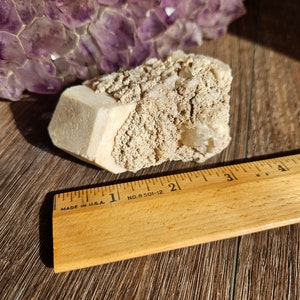 May include: A white, rough-textured crystal cluster with a large, smooth, white crystal in the center. The crystal cluster is sitting on a wooden surface next to a wooden ruler marked with inches. The ruler is labeled "MADE IN U.S.A." and "NO. R 501-12".