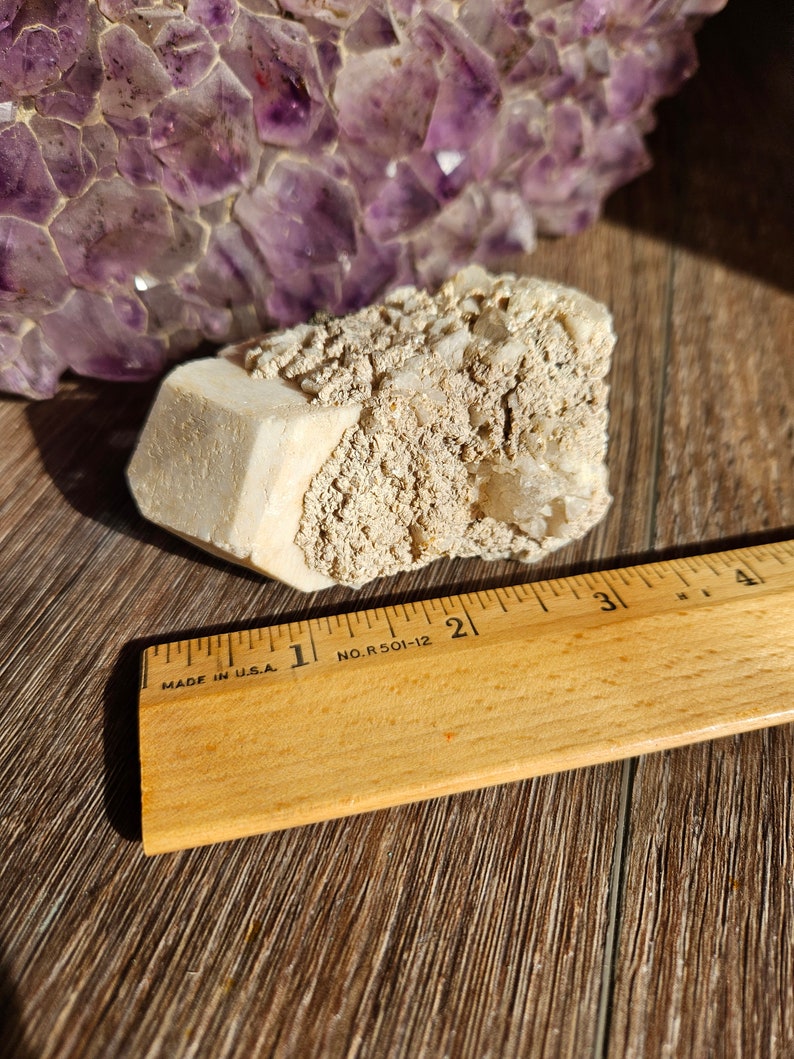 May include: A white, rough-textured crystal cluster with a large, smooth, white crystal in the center. The crystal cluster is sitting on a wooden surface next to a wooden ruler marked with inches. The ruler is labeled "MADE IN U.S.A." and "NO. R 501-12".