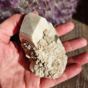 May include: A white crystal cluster with a rough, textured surface. The crystal has a pointed shape and is surrounded by smaller crystals.