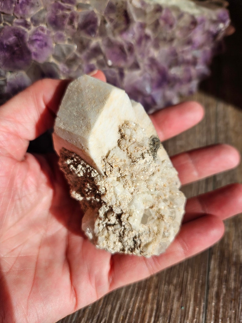 May include: A white crystal cluster with a rough, textured surface. The crystal has a pointed shape and is surrounded by smaller crystals.
