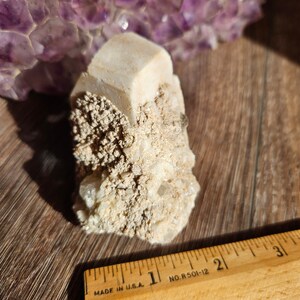 May include: A white, crystalline mineral cluster with a large, clear crystal in the center. The cluster is sitting on a wooden surface with a ruler next to it. The ruler is marked "MADE IN U.S.A." and "NO. R 501-12".