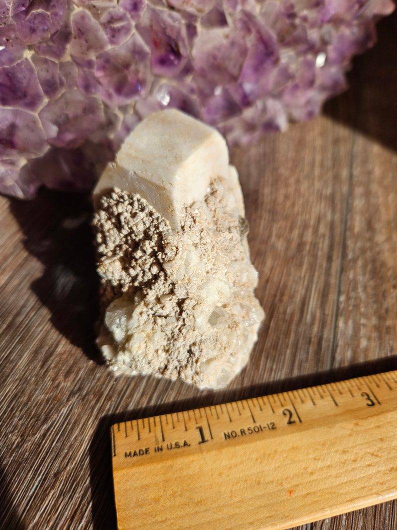 May include: A white, crystalline mineral cluster with a large, clear crystal in the center. The cluster is sitting on a wooden surface with a ruler next to it. The ruler is marked "MADE IN U.S.A." and "NO. R 501-12".