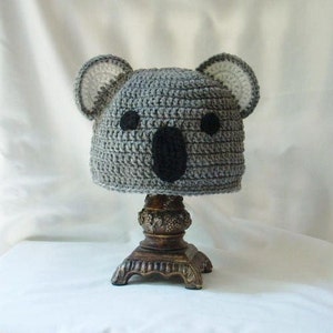 Op de afbeelding: Grijze gehaakte koala-muts met zwarte ogen en neus. De muts staat op een bruin voetstuk.