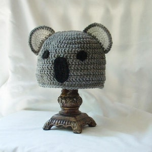 Op de afbeelding: Grijze gehaakte koala-muts met zwarte ogen en een witte gehaakte neus. De muts staat op een decoratieve standaard.