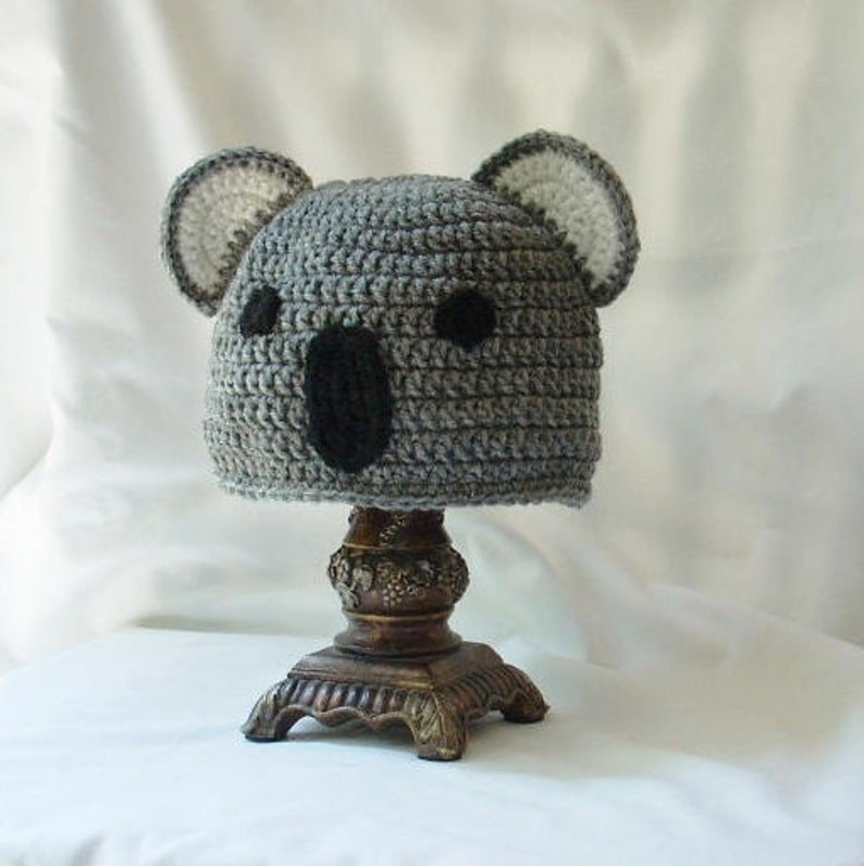 Op de afbeelding: Grijze gehaakte koala-muts met zwarte ogen en een witte gehaakte neus. De muts staat op een decoratieve standaard.