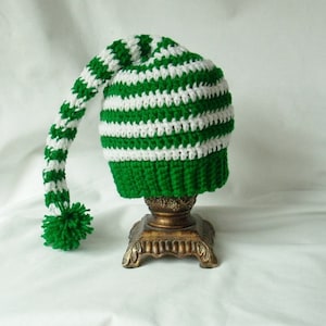 May include: A handmade crochet hat in green and white stripes, featuring a long, curved tip and a green pom-pom. The hat is displayed on a decorative, ornate stand, showcasing its whimsical design and craftsmanship.