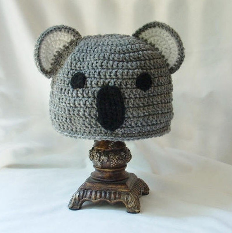 Op de afbeelding: Grijze gehaakte koala-muts met zwarte vilt ogen en neus. De muts wordt gepresenteerd op een houten standaard.