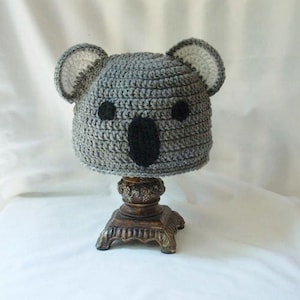Op de afbeelding: Grijze gehaakte koala-muts met zwarte ogen en neus. De muts staat op een houten standaard.
