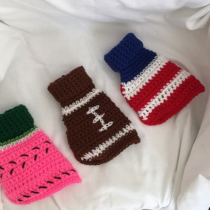 May include: Three crocheted mini-sweaters in the shape of a watermelon, football, and American flag. The watermelon is pink with green and white accents. The football is brown with white stripes. The American flag is red, white, and blue.