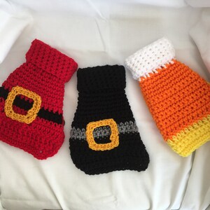 May include: Three crocheted dog sweaters in red, black, and orange and yellow. The red sweater has a black belt with a gold buckle. The black sweater has a gray and gold belt. The orange and yellow sweater has a white top.