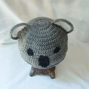Op de afbeelding: Grijze gehaakte koala-muts met zwarte ogen en neus. De muts staat op een houten standaard.