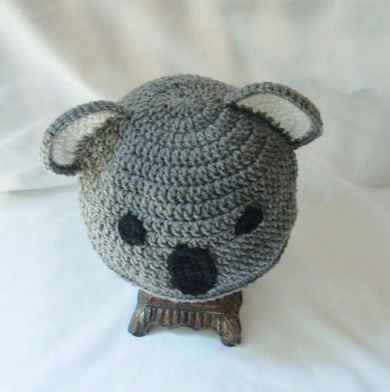 Op de afbeelding: Grijze gehaakte koala-muts met zwarte ogen en neus. De muts staat op een houten standaard.