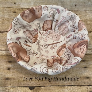 May include: Round, beige fabric bread basket with a repeating pattern of bread loaves, flowers, and sprigs. The text "Love You Big Handmade" is printed at the bottom. The basket is designed to hold bread.