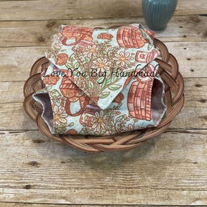 May include: A folded fabric item with a light blue background featuring a repeating pattern of orange picnic baskets, jars, and white daisies. The text Love You Big Handmade is printed across the center of the fabric. The item is placed inside a round woven basket.