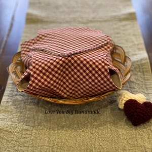 May include: A red and white gingham fabric basket liner with a brown wicker basket underneath. The text "Love You Big Handmade" is printed on the table below the basket. A small crocheted heart is on the table to the right of the basket.