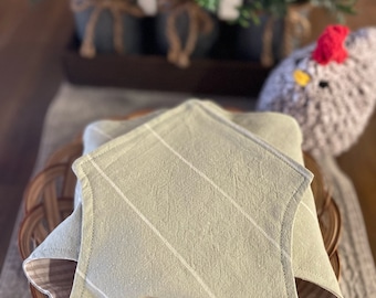 May include: A folded, sage green cloth napkin with white diagonal stripes, resting in a round, woven basket. The napkin is on a neutral-toned tablecloth. The text "Love You Big Handmade" is visible at the bottom.