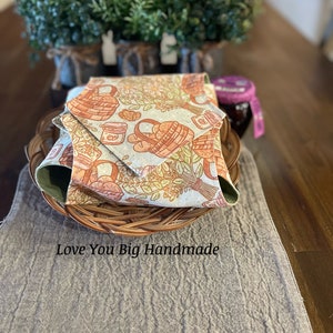 May include: A folded fabric with a basket and jar pattern in shades of brown, orange, and green. The fabric is in a wicker basket on a gray linen table runner. The text "Love You Big Handmade" is visible below the basket.