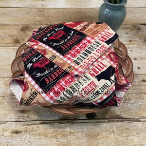 May include: A fabric bowl cover featuring a barbecue-themed print with red and white gingham patterns, black silhouettes of cows, and text including Roadhouse Grill, Bar-B-Q & Brew, and Smoked House. The fabric is folded in a woven basket.